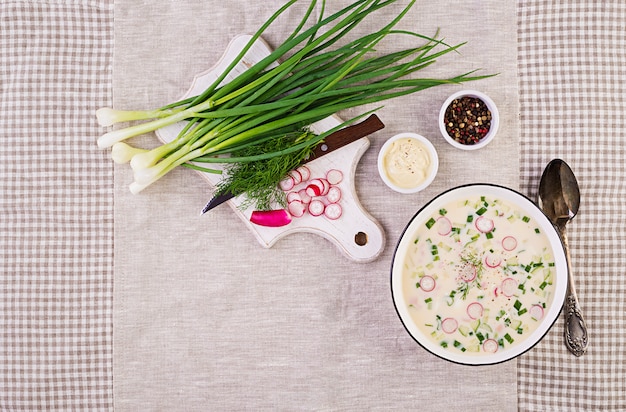 Sopa fría con pepinos frescos, rábanos, papas y salchichas con yogur en un tazón. Comida rusa tradicional - okroshka. Sopa fría de verano. Vista superior. Lay Flat