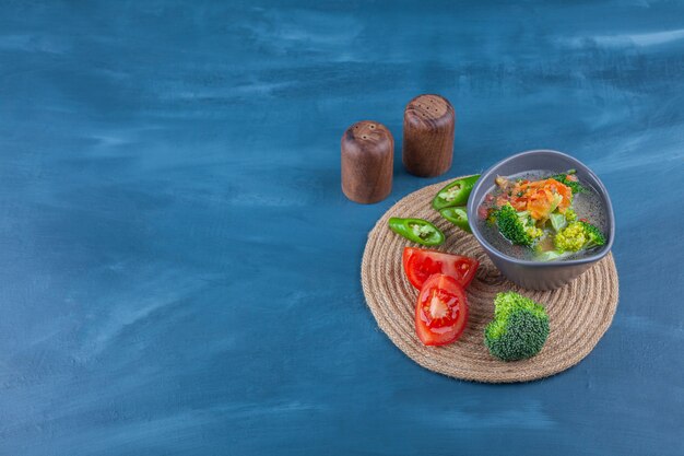 Sopa de pollo en un bol y verduras en rodajas sobre un salvamanteles, sobre la mesa azul.