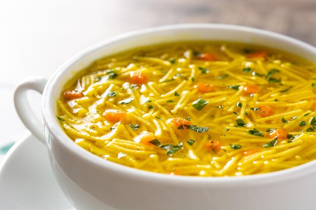 Sopa de pollo con fideos en un tazón blanco sobre una mesa de madera