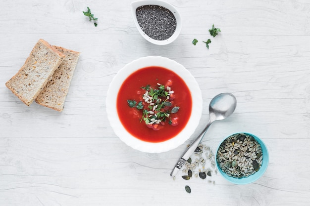 Sopa de tomate con chia y semillas de calabaza con una rebanada de pan en una mesa de madera blanca