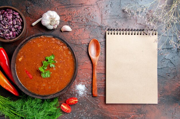 Sopa de tomate en un tazón marrón y diferentes especias ajo limón y cuaderno sobre mesa de colores mezclados