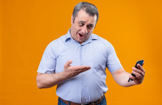 Sorprendente hombre en camisa a rayas azul hablando por videollamada con teléfono móvil mientras está de pie sobre un fondo naranja