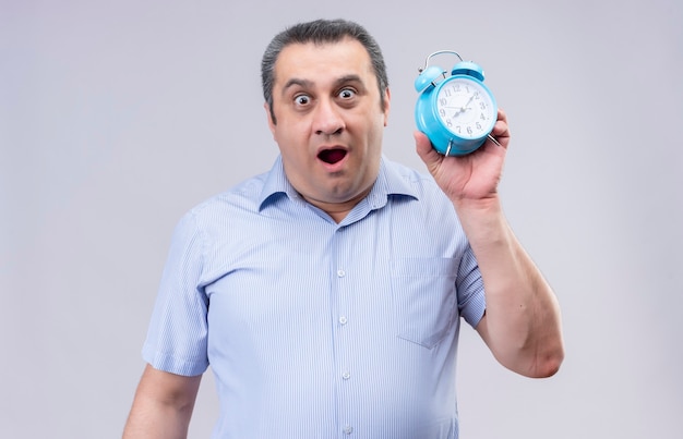 Sorprendente hombre de mediana edad vestido con camisa azul a rayas verticales sosteniendo reloj despertador azul mientras está de pie sobre un fondo blanco.