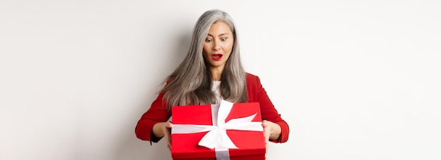 Foto gratuita sorprendida anciana asiática recibiendo un regalo el día de la madre sosteniendo una caja roja con un regalo y mirando un