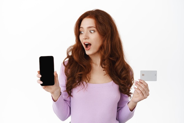 Sorprendida chica glamorosa muestra la aplicación de compras en la pantalla del teléfono inteligente y la tarjeta de crédito. La mujer miró fijamente su teléfono móvil con asombro, de pie contra el fondo blanco.