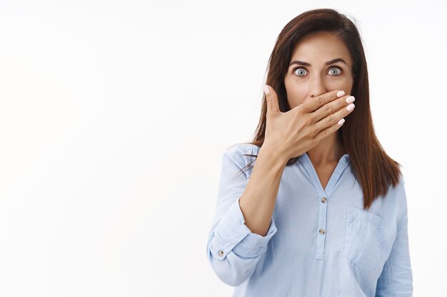 Sorprendida, divertida, mujer de mediana edad, morena, sorprendida, mira fijamente al frente, cubre la boca sin palabras, presiona los labios de la palma asombrados, chismes, escucha un rumor impresionante, posa una pared blanca