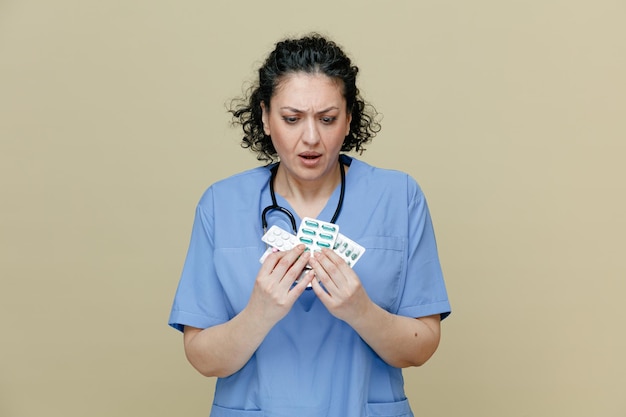 Sorprendida doctora de mediana edad con uniforme y estetoscopio alrededor del cuello sosteniendo paquetes de pastillas con ambas manos mirándolas aisladas en un fondo verde oliva