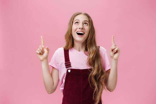 Sorprendida y encantada soñadora joven adolescente con elegantes monos de pana sonriendo ampliamente con una sonrisa emocionada mirando y apuntando hacia arriba disfrutando con alegría viendo el avión volando en el cielo sobre la pared rosa.