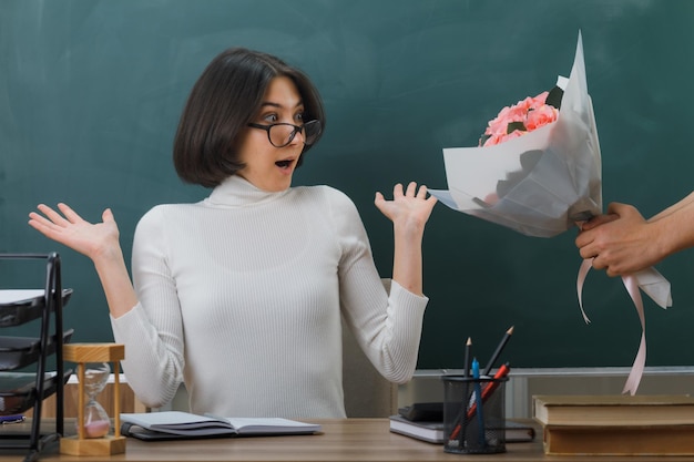 Foto gratuita sorprendida extendiendo las manos, la joven maestra sentada en el escritorio con herramientas escolares recibió un ramo de flores en el aula