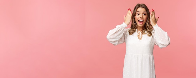 Sorprendida, feliz y emocionada niña de cumpleaños con vestido blanco, abre los ojos para ver un increíble regalo increíble de amigos en la fiesta de pie sobre un fondo rosa, se ve asombrada y alegre