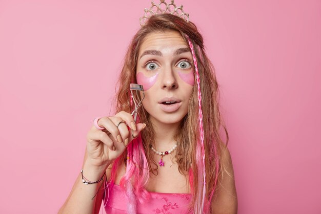 Sorprendida y hermosa mujer joven sostiene un rizador de pestañas, mira fijamente los ojos saltones, aplica parches de colágeno para eliminar las arrugas, ¿el maquillaje tiene el pelo largo, se prepara para la fecha aislada sobre fondo rosa?