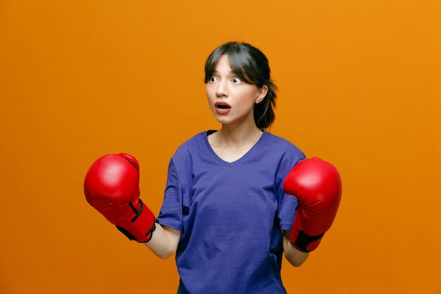 Sorprendida joven deportista con camiseta y guantes de boxeo mirando al costado manteniendo los puños en el aire aislados en el fondo naranja