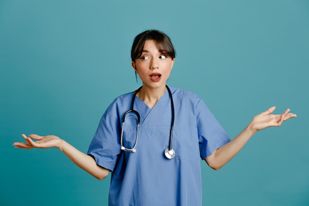 Sorprendida joven doctora vistiendo estetoscopio uniforme fith aislado sobre fondo azul.