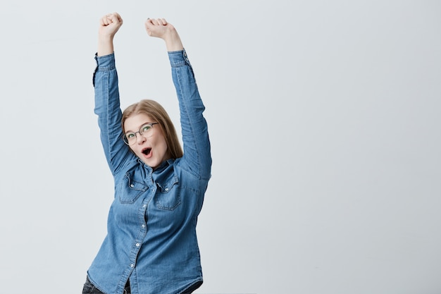 Foto gratuita sorprendida joven emocionada con cabello rubio lacio, gafas, con camisa de mezclilla levanta los brazos en el aire, celebrando su triunfo. feliz rubia joven regocijándose de su éxito.