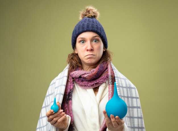 Sorprendida joven enferma vestida con túnica blanca y gorro de invierno con bufanda envuelta en cuadros con enemas aislados en verde oliva