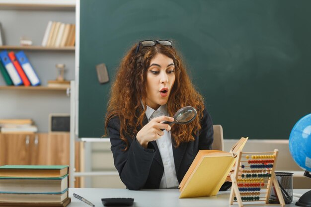 sorprendida joven maestra con anteojos leyendo un libro con lupa sentada en el escritorio con herramientas escolares en el aula