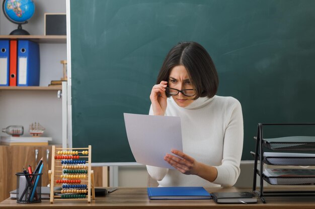 sorprendida joven maestra con anteojos sosteniendo y mirando papel sentado en el escritorio con herramientas escolares en el aula