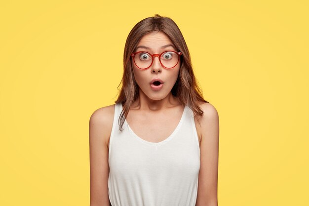 sorprendida joven morena con gafas posando contra la pared amarilla