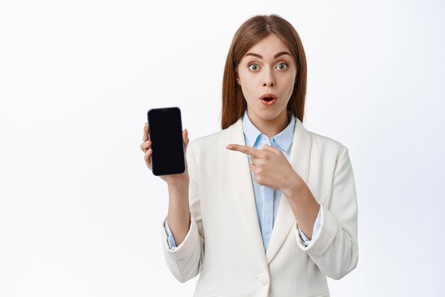 Sorprendida joven oficinista en traje de negocios jadea y los puntos que quedan en la pantalla del teléfono inteligente muestran una oferta promocional en línea de fondo blanco