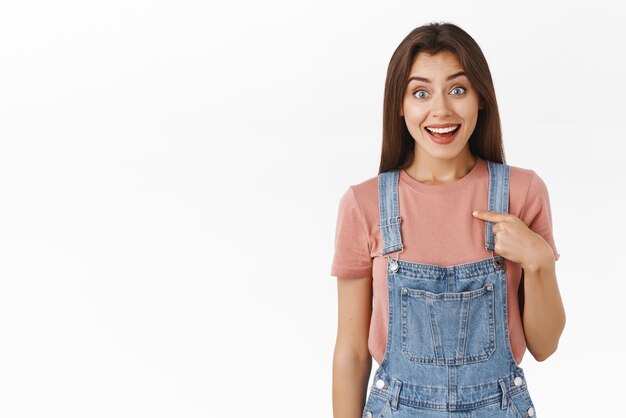 Sorprendida linda chica morena fascinada y feliz de ser elegida ganó algo increíble sonriendo felizmente apuntándose a sí misma y mirando la cámara radiante de alegría y alegría fondo blanco