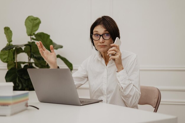 Sorprendida mujer adulta morena de piel clara con gafas y camisa usa un teléfono inteligente y una computadora portátil en una habitación luminosa
