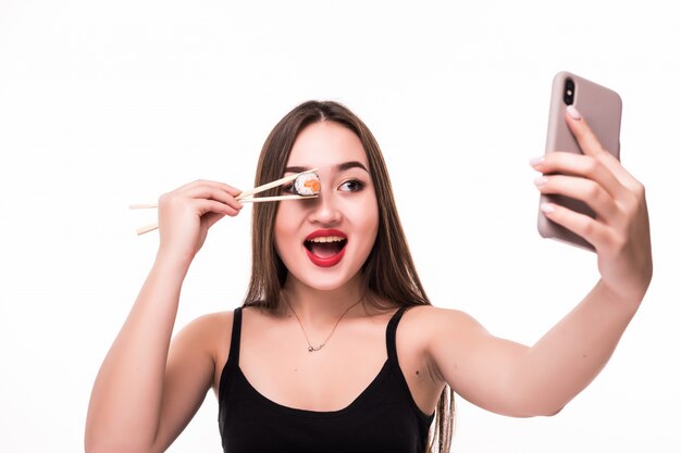 Sorprendida mujer asiática sonriente cubra su ojo con rollo de sushi y haga selfie en su teléfono