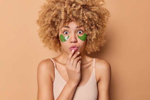 Sorprendida mujer de cabello rizado se ve maravillada ante la cámara que aplica parches de hidrogel verde debajo de los ojos para el rejuvenecimiento de la piel vestida con una camiseta informal aislada en un fondo beige Procedimientos matutinos en casa