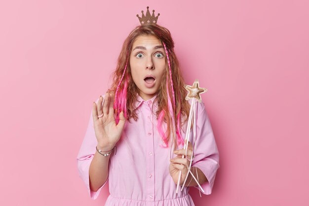 Sorprendida mujer joven asustada con el pelo largo mira impresionada a la cámara reacciona ante algo aterrador que sostiene una varita mágica que lleva una pequeña corona en la cabeza aislada sobre el fondo rosa del estudio Concepto Omg