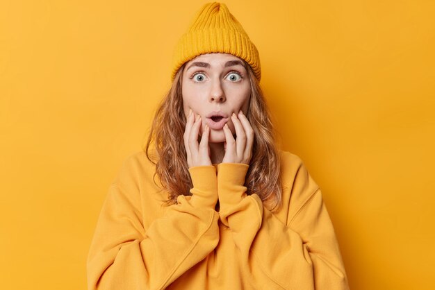 Sorprendida mujer joven de ojos azules mira fijamente a la cámara mantiene las manos en las mejillas con la boca abierta usa una sudadera informal y el sombrero reacciona ante algo aterrador aislado sobre el concepto de fondo amarillo Omg