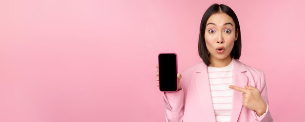 Sorprendida mujer de negocios asiática entusiasta que muestra la interfaz de la aplicación del teléfono inteligente de la pantalla del teléfono móvil de pie contra el fondo rosa