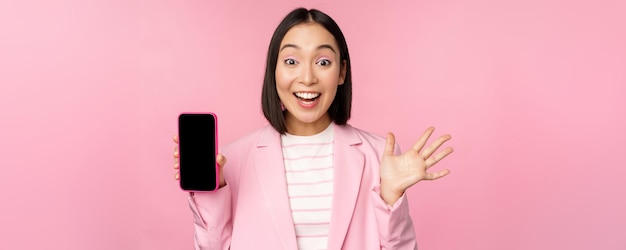 Sorprendida mujer de negocios asiática entusiasta que muestra la interfaz de la aplicación del teléfono inteligente de la pantalla del teléfono móvil de pie contra el fondo rosa