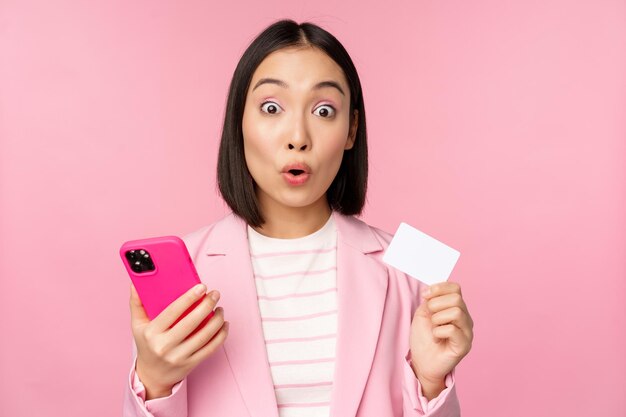 Sorprendida mujer de negocios asiática con traje que muestra compras en línea con tarjeta de crédito y pedidos de teléfonos móviles con un teléfono inteligente de pie sobre fondo rosa Concepto de publicidad