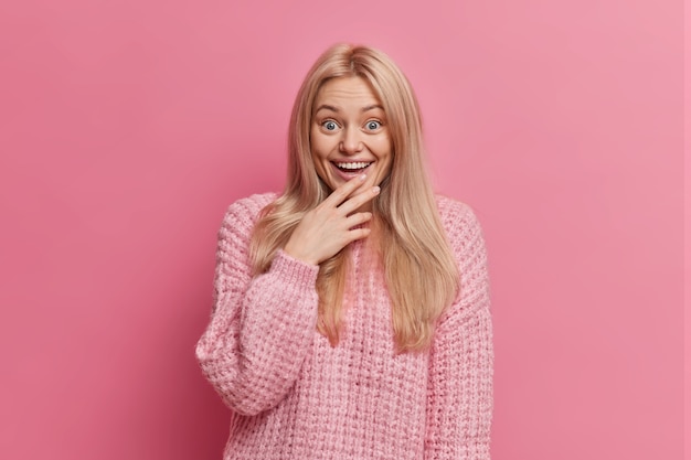 Sorprendida mujer rubia divertida mira algo maravilloso con una amplia sonrisa pierde el habla del asombro vestida con un cálido suéter de invierno