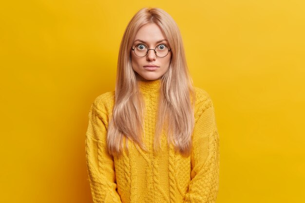 sorprendida mujer rubia se encuentra sin palabras en el interior lleva gafas redondas transparentes vestidas con un suéter amarillo plantea interior. Mujer europea impresionado sorprendido mira fijamente los ojos saltones.