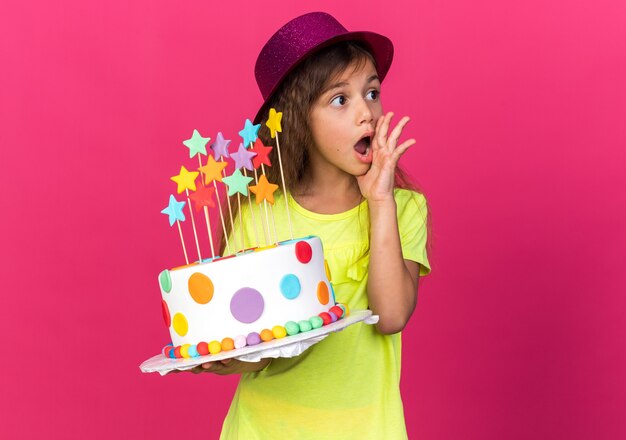 Sorprendida niña caucásica con sombrero de fiesta púrpura poniendo la mano en la cara y sosteniendo la torta de cumpleaños mirando al lado aislado en la pared rosa con espacio de copia