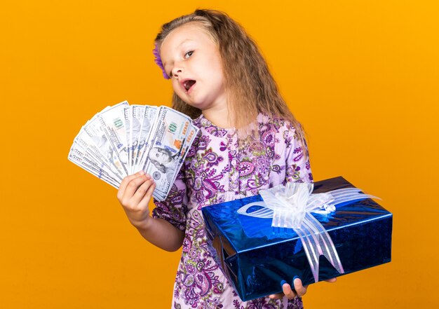 Sorprendida niña rubia con caja de regalo y dinero aislado en la pared naranja con espacio de copia