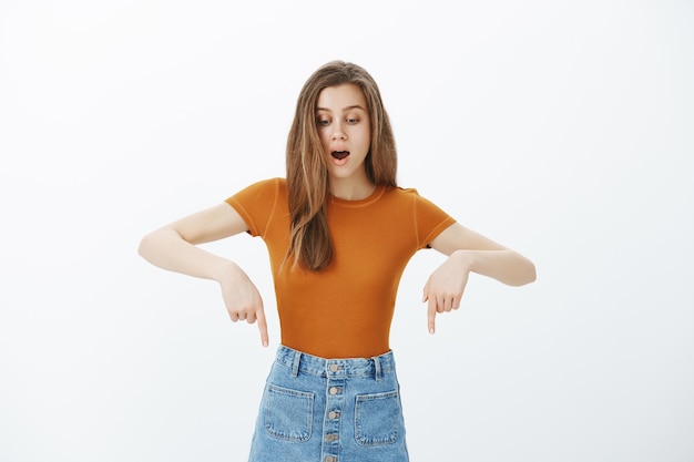 Foto gratuita sorprendida y sorprendida mujer atractiva señalando con el dedo hacia abajo, jadeando asombrado