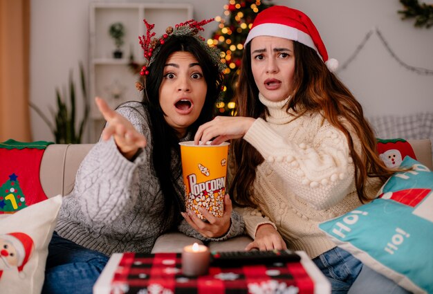 Sorprendidas chicas guapas con gorro de Papá Noel y corona de acebo comen palomitas de maíz sentadas en sillones y disfrutando de la Navidad en casa