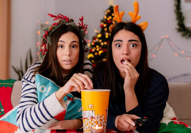 Sorprendidas chicas jóvenes con corona de acebo y diadema de renos comen palomitas de maíz viendo la televisión sentadas en sillones y disfrutando de la Navidad en casa