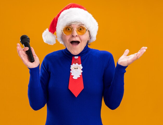 Sorprendido anciana en gafas de sol con gorro de Papá Noel y corbata de santa sosteniendo micrófono aislado sobre fondo naranja con espacio de copia