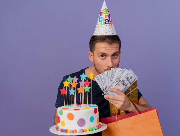 Sorprendido apuesto hombre caucásico con gorro de cumpleaños tiene caja de regalo de bolsa de papel de tarta de cumpleaños y dinero aislado sobre fondo púrpura con espacio de copia