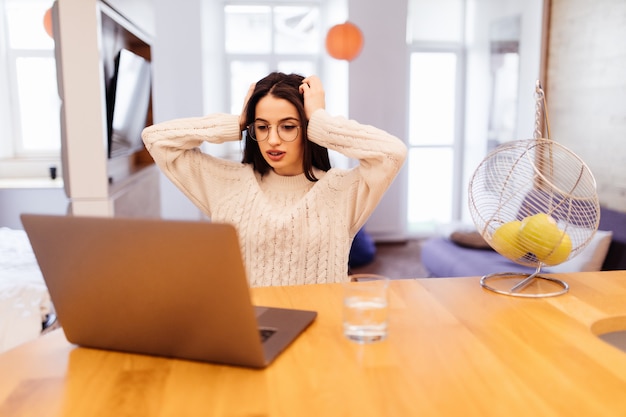 Sorprendido Bastante joven está sentada en la cocina y trabajando en su computadora portátil y teléfono móvil