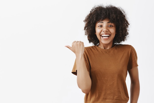 sorprendido complacido e impresionado feliz atractiva mujer de piel oscura en t-shrit marrón con cabello rizado apuntando hacia la izquierda con el pulgar y sonriendo explicando lo increíble que sucedió