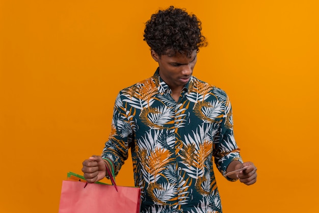 Sorprendido y confundido joven apuesto hombre de piel oscura con cabello rizado en hojas camisa impresa sosteniendo bolsas de compras y mirando extrañamente una tarjeta de crédito mientras está parado en un centrico naranja