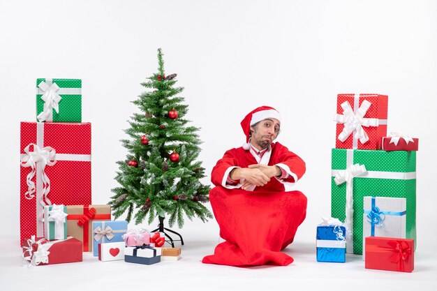 Sorprendido emocional emocionado joven adulto vestido como Papá Noel con regalos y árbol de Navidad decorado sentado en el suelo sobre fondo blanco.