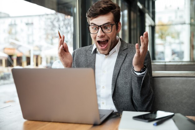 Sorprendido empresario gritando en anteojos sentado junto a la mesa en la cafetería y mirando la computadora portátil