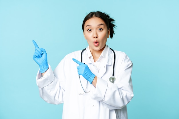 Sorprendido y entusiasta médico asiático, trabajadora médica señalando con el dedo en la esquina superior izquierda, mostrando disocunt, de pie sobre fondo azul