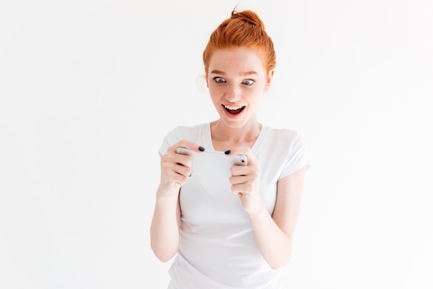 Sorprendido feliz mujer de jengibre en camiseta jugando en su teléfono inteligente