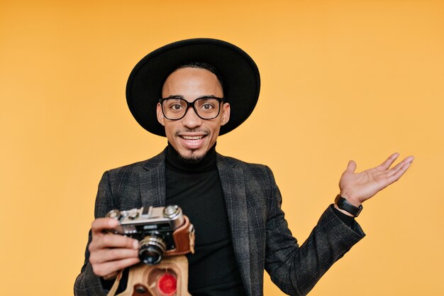 sorprendido fotógrafo masculino con sombrero negro. Foto interior de joven africano con cámara aislada en pared amarilla.