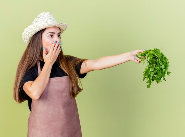 Sorprendido hermosa niña jardinero en uniforme con sombrero de jardinería sosteniendo cilantro en el costado y boca cubierta con la mano aislada en verde oliva